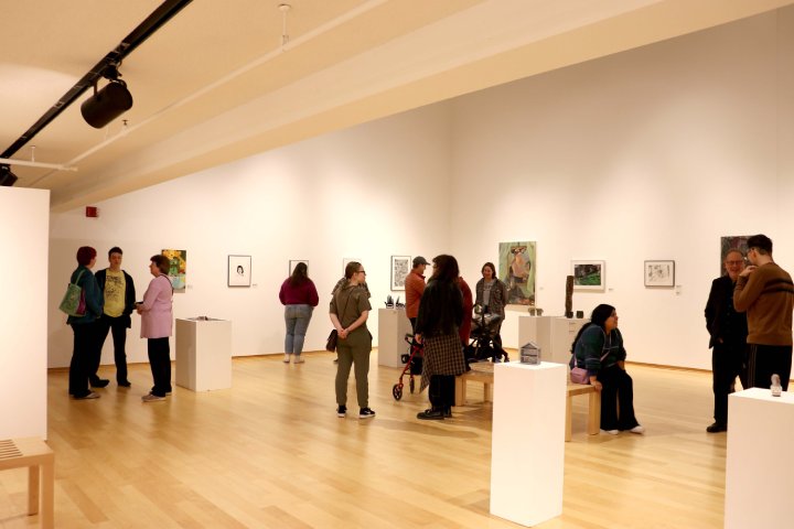 People gather and converse inside an art gallery, with paintings and sculptures displayed on the walls and pedestals. The space has light wooden floors and bright, even lighting, emphasizing the artwork.